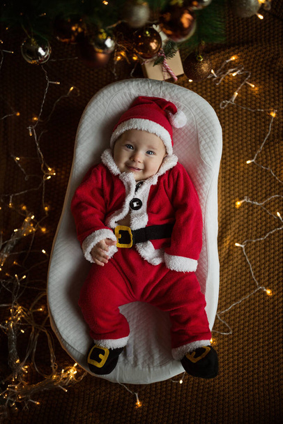 Closeup portrait of newborn baby. Cute Caucasian baby girl 4-5 months old in Santa costume lying on knitted cozy blanket in cocoon near decorated fir tree. Merry Christmas xmas and happy new year 2022 - Photo, image