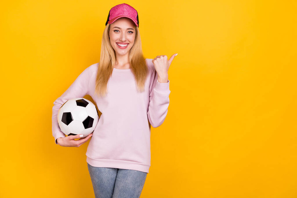 Photo of pretty adorable woman dressed pink pullover headwear holding ball pointing thumb empty space isolated yellow color background - Fotografie, Obrázek