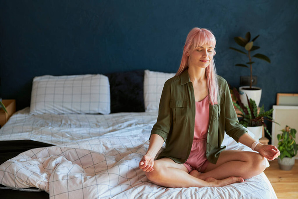 Positive young woman in green shirt and pajamas meditates sitting in lotus pose on large bed - Фото, зображення