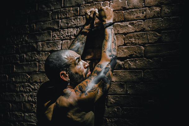 Ένα φαλακρό μέλος συμμορίας με τατουάζ στέκεται απέναντι από τον τοίχο με τα χέρια του πάνω σε χειροπέδες. Σύλληψη επικίνδυνου εγκληματία. - Φωτογραφία, εικόνα