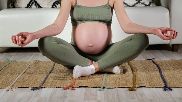 Pregnant lady sits in lotus position on mat against sofa - Photo, Image