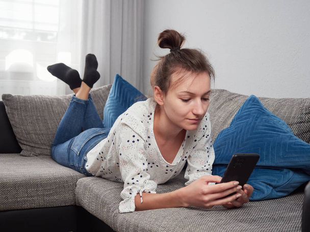 Una mujer joven está acostada en el sofá de la sala de estar y está usando un teléfono inteligente. Chica relajada navegando por Internet en un teléfono inteligente. - Foto, Imagen