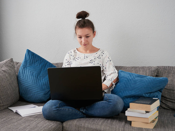 Genç bir kadın kanepede oturuyor ve bir dizüstü bilgisayar kullanıyor. Kitaplar ve defterler onun yanında duruyor. Öğrenci evde öğreniyor. E-öğrenme. - Fotoğraf, Görsel