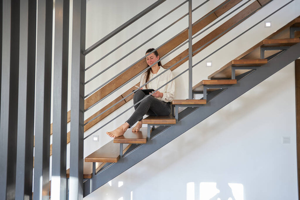 Woman working from home in her living room while sitting on staircase. Modern metal stairway in interior design - Foto, immagini