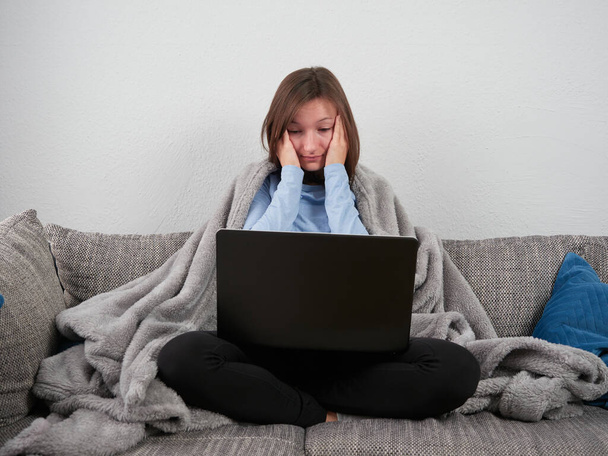 Eine junge, müde Frau sitzt in eine weiche Decke gehüllt auf dem heimischen Sofa und benutzt einen Laptop. Ein gelangweiltes Mädchen hält ihre Hände auf dem Gesicht. - Foto, Bild