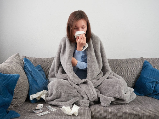 A sick young woman wrapped in a soft blanket is sitting on the couch at home. A girl with a runny nose is blowing her nose into a handkerchief. There are tablets in front of her. - Foto, Imagem