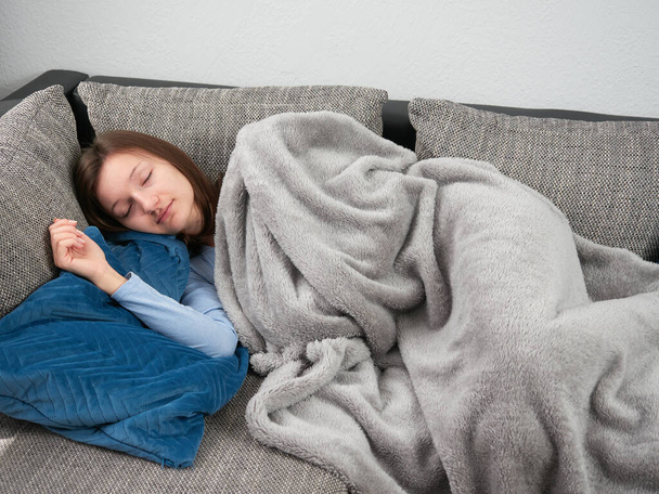 Όμορφη νεαρή γυναίκα τυλιγμένη σε μια απαλή κουβέρτα κοιμάται στον καναπέ στο σπίτι. Το κορίτσι παίρνει έναν απογευματινό υπνάκο. - Φωτογραφία, εικόνα