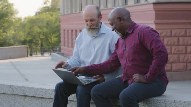 Dva legrační starší muži sedí venku a používají notebook, hledají štěnici, dostávají negativní zprávy. Afričtí a evropští důchodci mají problémy s online aplikacemi, moderními technologiemi - Záběry, video