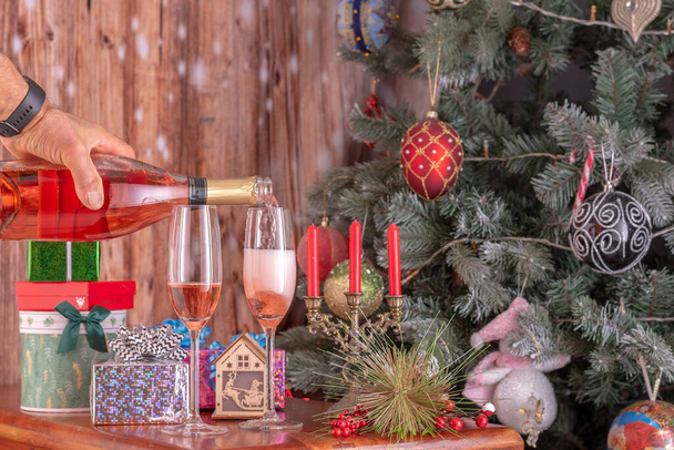 Herrenhände, die Champagner aus einer Flasche in Champagner gießen, flattern vor dem Hintergrund eines Neujahrsbaums und überreichen Geschenke. - Foto, Bild