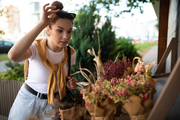 mujer joven caucásica feliz elige flores en maceta para comprar en el puesto de jardín al aire libre - Foto, Imagen