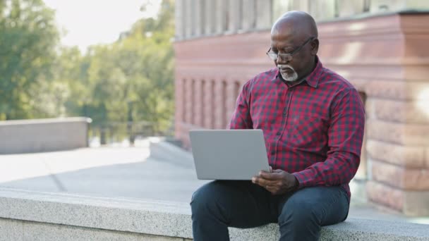 Ernstige gefocuste senior zwarte persoon met behulp van laptop, Afro-Amerikaanse volwassen man communiceren op sociale netwerken online, oudere man het schrijven van e-mails, op afstand werken op de computer in het openbaar - Video