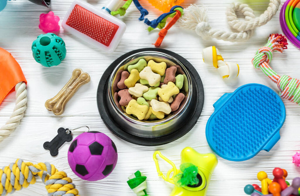 Different multicolored pet care accessories: bowl, bones, balls, snacks on white wooden background. Rubber and textile toys for dogs. Top view, flat lay. Copy space - Foto, Bild