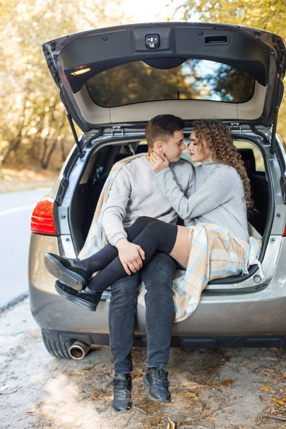 The guy and the girl are hugging in the trunk of the car. Autumn forest. Concept for love, happy family, travel and weekend. Vertical. - 写真・画像