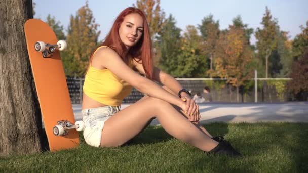 Giovane bella donna con uno skateboard si siede nel parco sull'erba. Ritratto di una bella donna rossa attiva all'aperto. Nuova generazione - Filmati, video