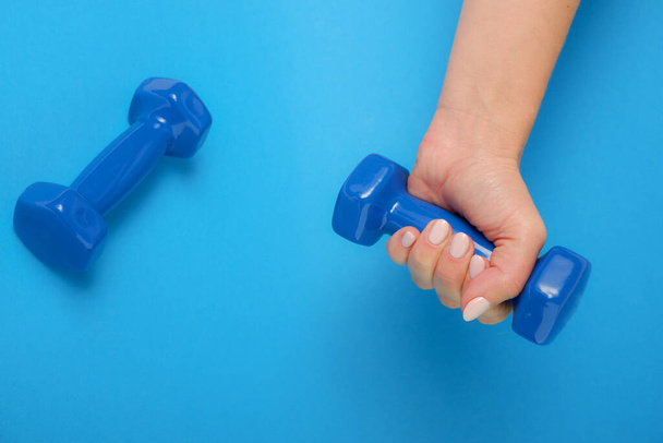 Concepto fitness Una mano femenina sostiene una mancuerna de plástico azul. Fondo azul. - Foto, imagen
