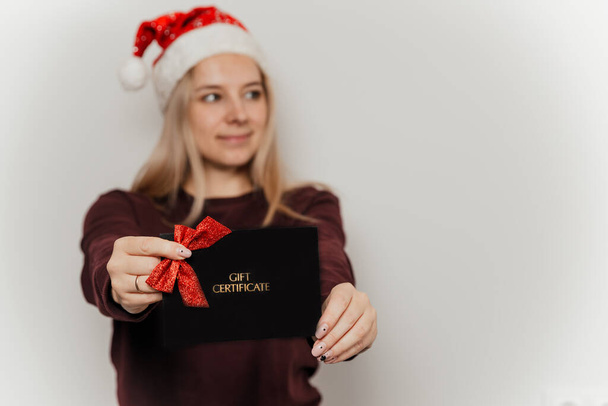 Woman in a burgundy sweater and red santa hat holding black gift certificate on a gray background - Photo, Image