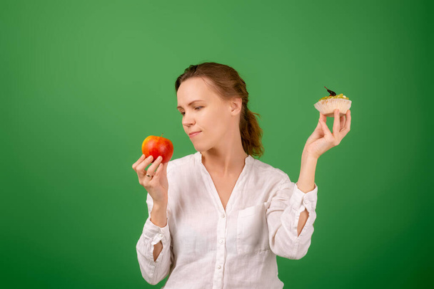 Middle-aged woman with apple and cake on a green background. Looks at the apple. The concept of choosing the right healthy food. - Photo, Image