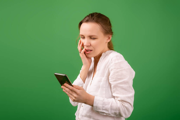 A shocked middle-aged woman in a white shirt looks at the phone on a green background. - 写真・画像