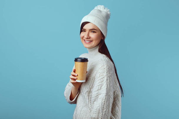Šťastná dívka v bílém klobouku a bílém teplém svetru, drží odnášející šálek kávy - Fotografie, Obrázek