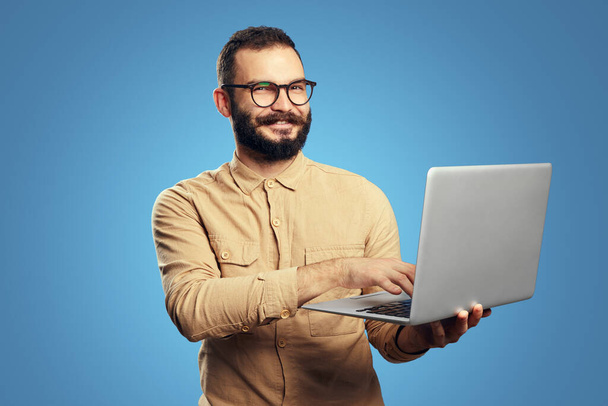 Homme portant une chemise beige et des lunettes souriant tout en tenant et en utilisant un ordinateur portable - Photo, image