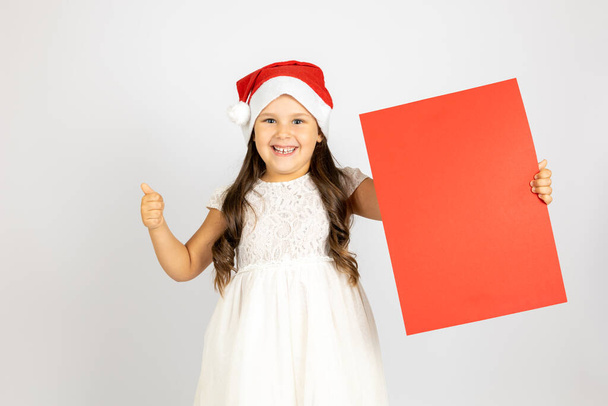 portrait of joyful, cheerful girl in white dress and Santa Claus hat gives thumbs up and holds red paper, isolated on white background - Фото, зображення