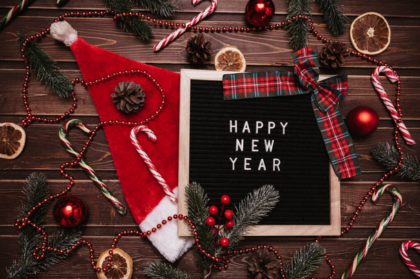 Happy New Year inscription on a letter board in New Year decorations, Santa hat, fir branches, cones, balls, caramel canes. Top view. Christmas card. - 写真・画像