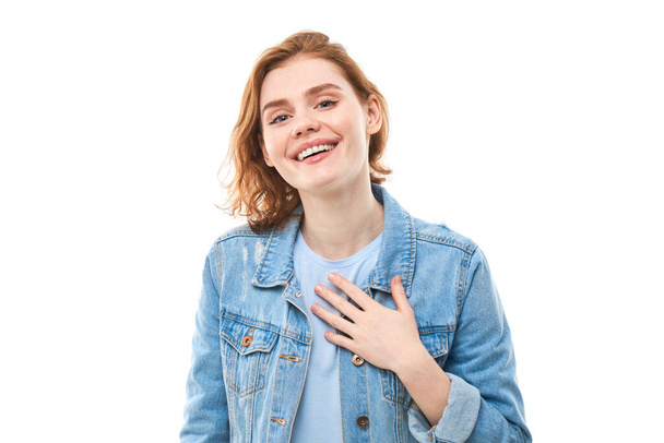 Lovely good-natured red-haired girl student in a blue denim jacket with a charming smile and a friendly expression, holding hands on her heart, wanting to show her love and sympathy. On a white background - Zdjęcie, obraz