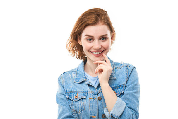 A girl student with red hair happily thinks and looks at the camera while holding her finger near her chin. Pensive thoughtful woman in a blue denim jacket on a white background. Copy space - Foto, imagen