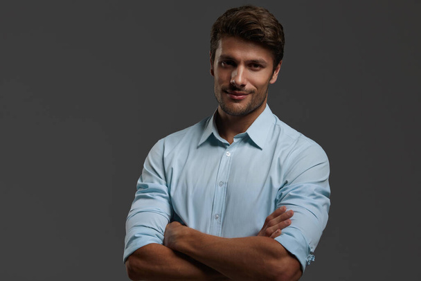 Young beautiful smiling european businessman with crossed arms. Bearded man with dark hair wear blue shirt and looking at camera. Isolated on dark gray background. Studio shoot. Copy space - Фото, изображение