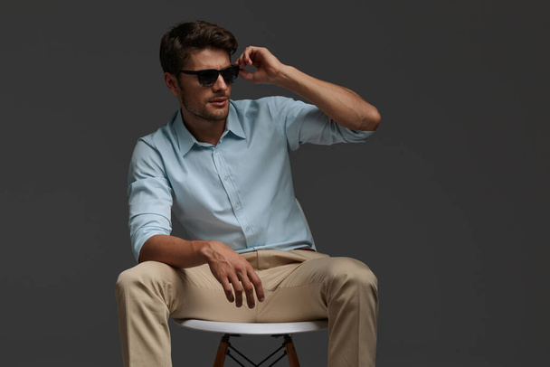 Young beautiful european businessman sitting on chair. Bearded man with dark hair wear shirt with trousers and glasses. Isolated on dark gray background. Studio shoot. Copy space - Foto, immagini