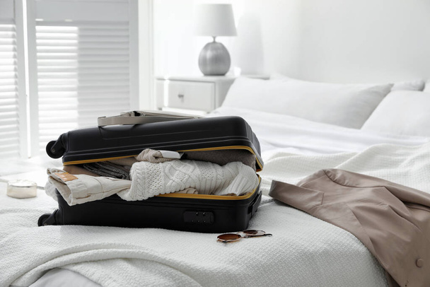 Open suitcase full of clothes, jacket and fashionable accessories on bed in room - Fotoğraf, Görsel