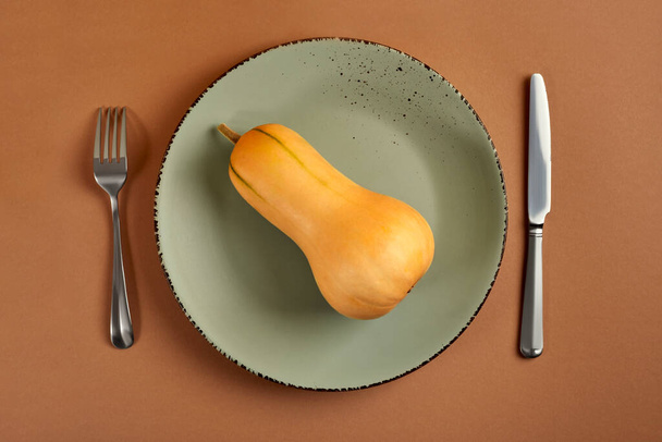 La citrouille repose sur une assiette verte. Une fourchette et un couteau sont à côté de l'assiette. La plaque se tient sur un fond brun. Vue d'en haut, plan plat. - Photo, image
