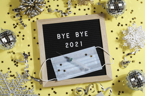 tablero de letras negro con texto BYE BYE 2021 con decoración, jeringa y máscara sobre fondo amarillo.  - Foto, Imagen