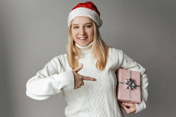 Beyaz süveterli ve kırmızı Noel Baba şapkalı bir kadın elinde gri bir arka planda pembe bir hediye tutuyor. - Fotoğraf, Görsel