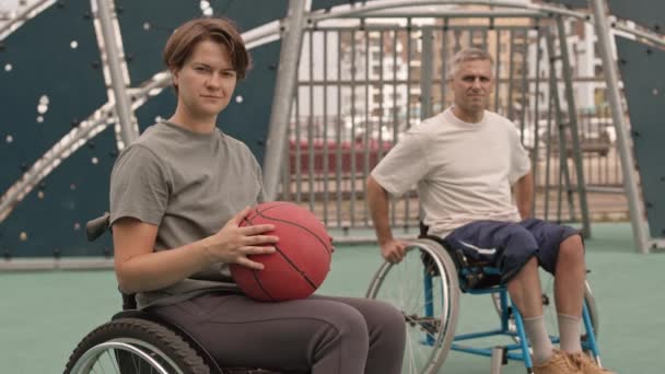 Μέτριο μήκος της νεαρής καυκάσιας γυναίκας με σωματική αναπηρία κρατώντας μπάσκετ, θολή γκριζομάλλης άνδρας σε αναπηρική καρέκλα στο παρασκήνιο, οι άνθρωποι κοιτάζουν στην κάμερα, ποζάρουν σε υπαίθριο γήπεδο - Πλάνα, βίντεο