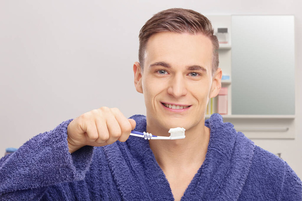 Young man in a bathrobe brushing his teeth in front of a mirror in a bathroom - Photo, Image
