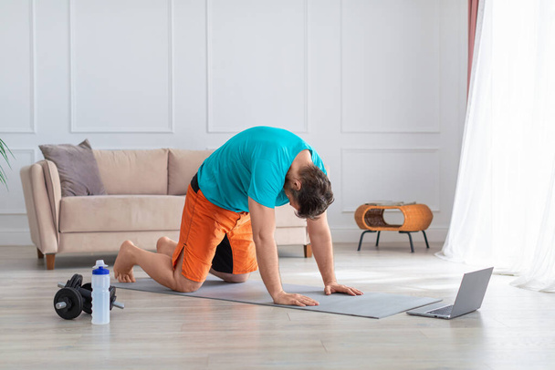 Онлайн тренировки на дому - мужчина среднего возраста во время тренировки йоги дома. Он вытягивает спину перед монитором ноутбука. - Фото, изображение