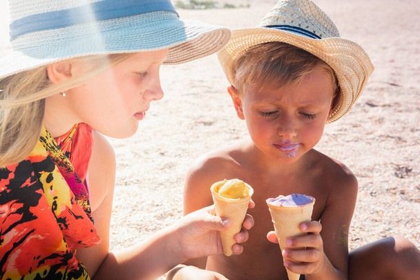 relaxing in the air, a blond boy and a blond girl in hats sitting on the beach, on the sand and eating or tasting ice cream with a small depth of field and copy space. Vacation concept   - Photo, Image