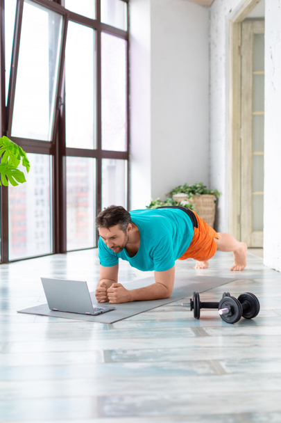 Verticale foto van een man van middelbare leeftijd voor de laptop monitor doet de plank oefening. Hij volgt de begeleiding van zijn online coach. - Foto, afbeelding