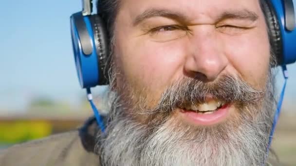 Brodaty mężczyzna w średnim wieku ze słuchawkami słucha muzyki, śpiewa i kiwa głową w rytm muzyki. Zbliżenie twarzy. - Materiał filmowy, wideo