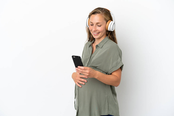 Mujer joven caucásica aislada sobre fondo blanco embarazada y escuchando música con un móvil - Foto, imagen