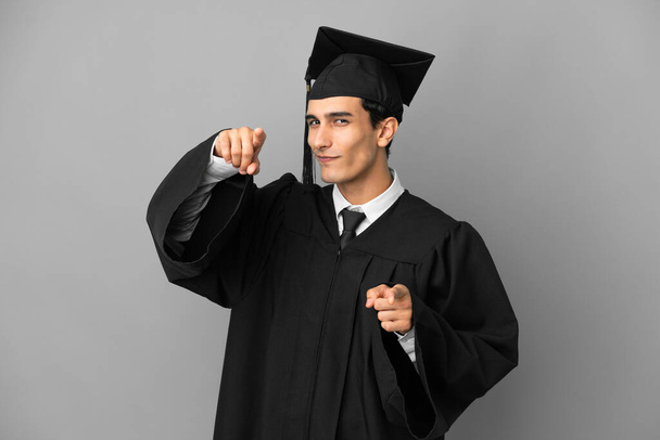 Joven graduada universitaria argentina aislada sobre fondo gris te señala con el dedo mientras sonríe - Foto, imagen