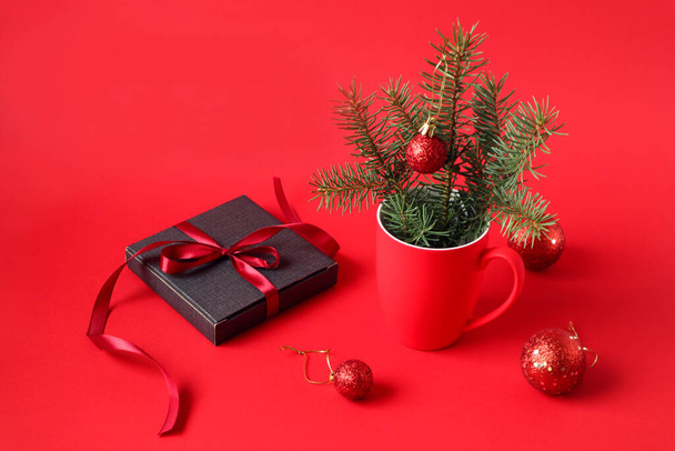 Postcard: a gift in a gray package, tied with a red ribbon, a red cup with a fir branch, Christmas balls scattered on a red background - Foto, immagini