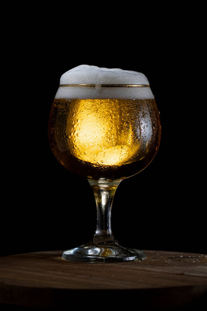 Beer glass with moisture drops on glass and white foam cap on black background - Фото, зображення