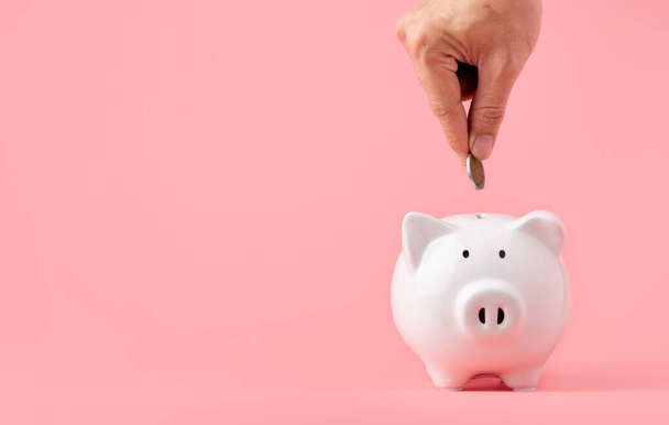 Money saving and investment concept. Hand putting coin to piggy bank on pink background with copy space - Fotó, kép