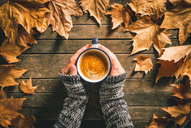 Kafkas bir erkeğin elleri, etrafında sonbahar yaprakları olan eski ahşap bir masanın üzerinde sıcak kahve fincanı tutuyor. Ev gibi hisset ve rahatla. - Fotoğraf, Görsel