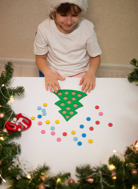 dětská dívka v bílém tričku s novoroční čepicí na hlavě stříhá originální stromeček na vánoční stromeček. koncepce vánočních tradic. vývoj jemných motorických dovedností Montessori na Vánoce - Fotografie, Obrázek