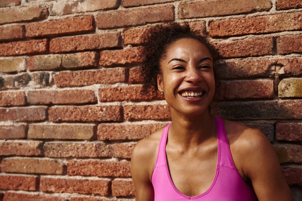 Happy young sportswoman laughing cheerfully while standing against a brick wall outdoors during the day - Photo, image