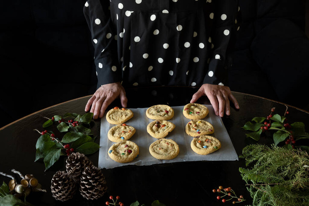Unrecognizable woman leaving trays of Christmas cookies on table, black background with copy space. - Photo, Image