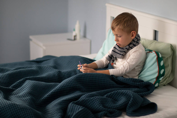Egy sálas kisfiú beteg és fekszik az ágyban, egy hőmérőt tartva a kezében. Szezonális hideg koncepció.  - Fotó, kép